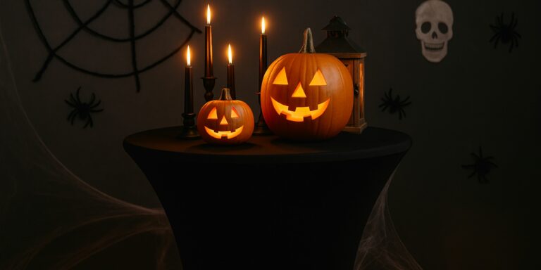 Schwarze Stehtischhusse mit zwei leuchtenden geschnitzten Kürbissen, schwarzen Kerzenhaltern, Laterne und Spinnennetz-Deko vor dunklem Hintergrund mit Spinnen und Totenkopf.